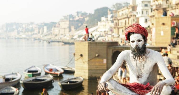 Asbestoven Sadhu in rode tulband poseert naast de Ganges met boten en Varanasi ghats die vervagen in de nevel.