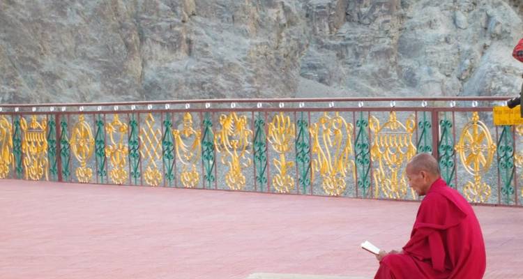 Mönch in roten Gewändern liest ruhig an einem bunt bemalten Geländer mit felsiger Klippe dahinter