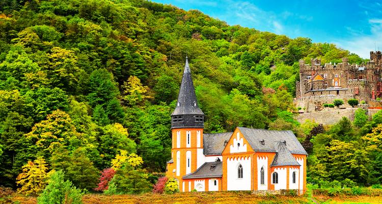 Église blanche et orange avec un haut clocher et un château sur la colline niché dans la forêt luxuriante des gorges du Rhin.