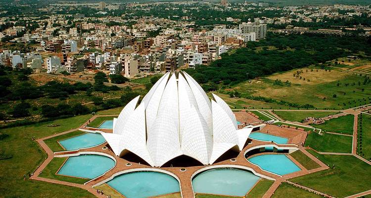 Luchtfoto van de witte bloembladachtige structuur van de Lotus Tempel omringd door vijvers en het stadslandschap erachter.