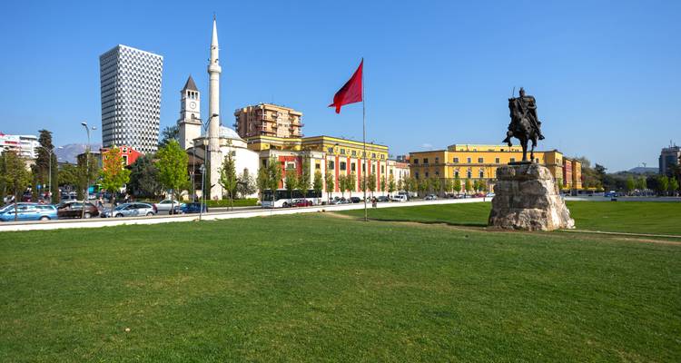 Plaza Skanderbeg en Tirana con estatua ecuestre, mezquita, torre del reloj, rascacielos moderno y bandera albanesa.