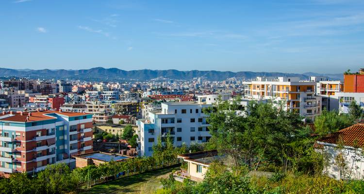 Vista panorámica del paisaje urbano de los barrios residenciales de Tirana con montañas distantes bajo un cielo azul despejado.