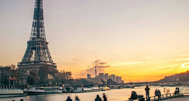 La Torre Eiffel se alza sobre el Sena al atardecer con siluetas difusas de transeúntes a lo largo de la ribera