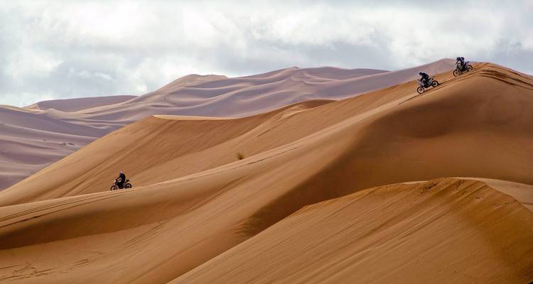 Motorcyclists ride along steep ridges of towering Sahara sand dunes.