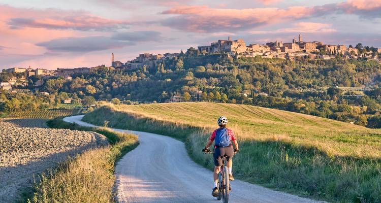 Cyclist rides along a winding dirt road toward a hilltop Tuscan village at sunset with pink clouds overhead.