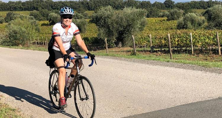 Smiling cyclist on a road bordered by vineyards and olive trees.