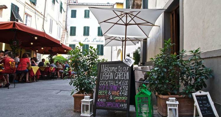 Charming Italian alleyway café with chalkboard menu, potted plants and diners seated under umbrellas.