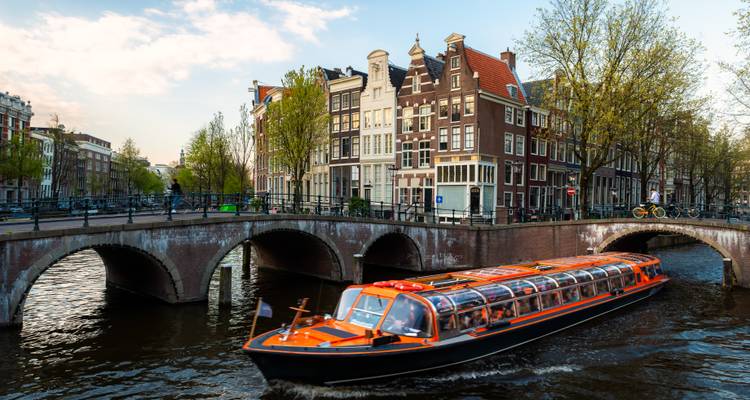 Orange glass-roof canal cruiser passing under a brick bridge between tall Amsterdam canal houses in warm late-afternoon light.