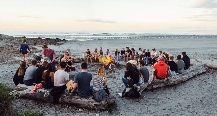 Een grote groep verzamelt zich rond een strandvuur bij schemering met golven op de achtergrond.