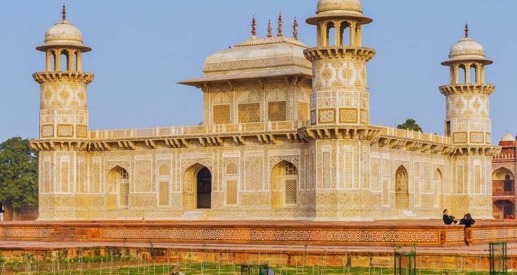 Itimad-ud-Daulahs kunstvoll verziertes Marmormausoleum in Agra mit zwei Besuchern, die auf seinem roten Sandsteinsockel sitzen.