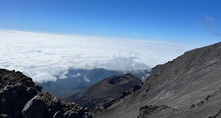 Borde del cráter y laderas volcánicas que descienden hacia un mar de nubes bajo un cielo azul despejado.