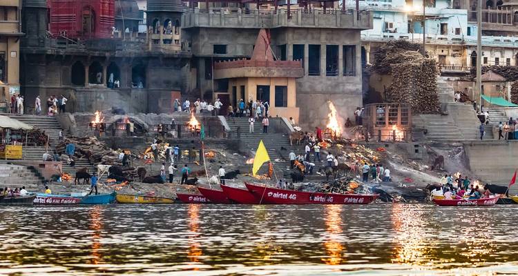 Lodernde Verbrennungsghaats und farbenfrohe Boote säumen die Ganges-Uferpromenade in Varanasi in der Dämmerung.