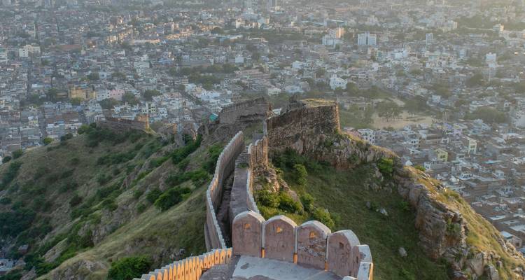 Luftaufnahme einer langen Steinfortmauer, die sich über grüne Hügel oberhalb der ausgedehnten Stadt Jaipur schlängelt.