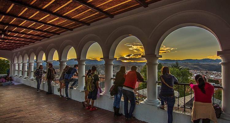 Visitors gather along an arched colonial balcony in Sucre watching the sun set over distant hills.