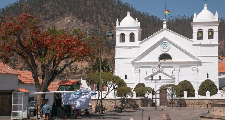 Small white colonial church framed by red-flowering tree with Andean hills in the background.