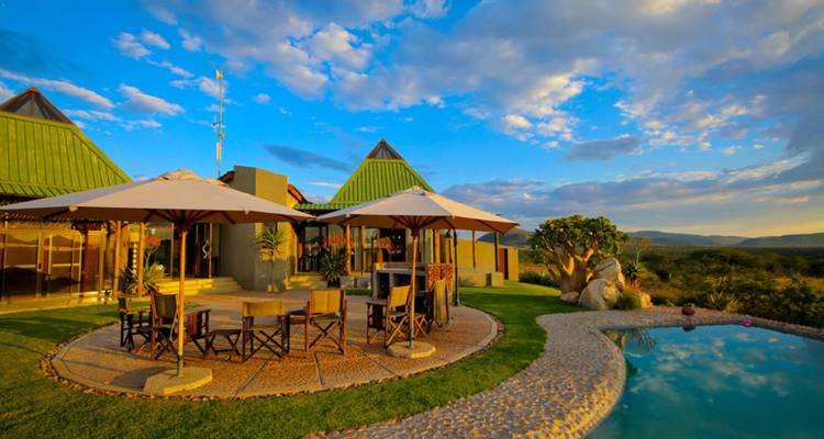 Stylish safari lodge with umbrellas, outdoor seating and a small swimming pool against a dramatic golden-hour sky and distant hills.