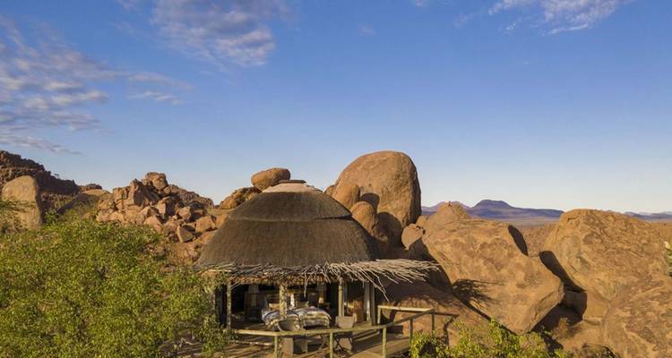 Thatched-roof hut nestled among giant reddish boulders overlooking open plains at dawn.