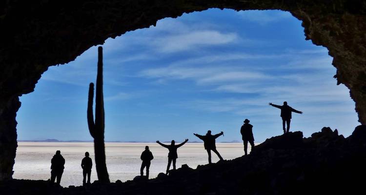 Silhouetted figures with outstretched arms stand in a cave opening overlooking the Uyuni salt flat.
