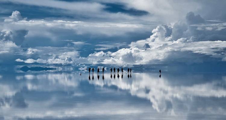 Dozens of tourists appear as tiny reflections on the mirror-like flooded Uyuni salt flats under dramatic clouds.