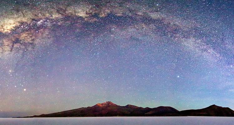 Panoramic arch of the Milky Way over a dark volcanic island and the Uyuni salt flat at night.