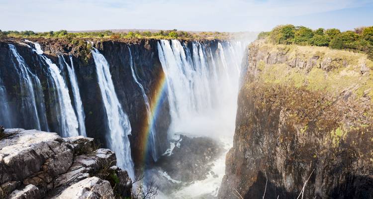 Powerful Victoria Falls plunges into a narrow gorge with a rainbow in the mist and lush green rim.