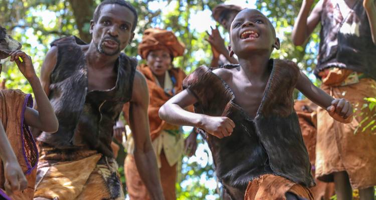 Traditional performers wearing animal skin costumes dance energetically in a forest setting.