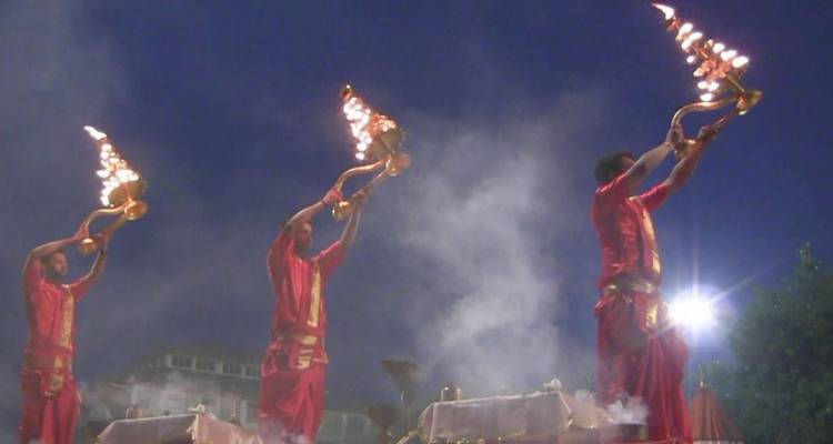 Drie priesters in rode gewaden heffen vlammende koperen lampen omhoog te midden van rituele rook tijdens de avond Ganga Aarti.