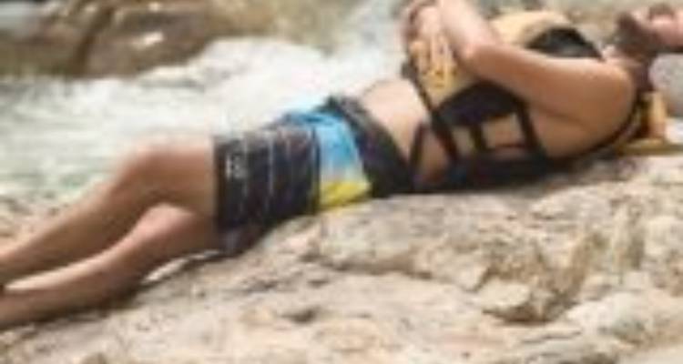 Blurry image of a rafter resting on a riverside boulder in swim shorts and life vest.