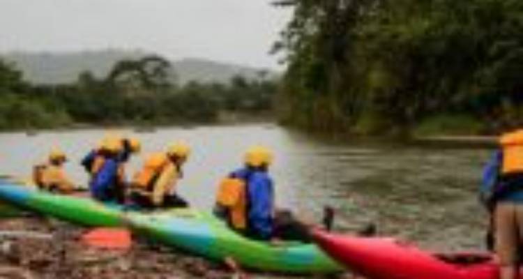 Blurry kayakers in helmets prepare to launch from a muddy riverbank on a cloudy day.