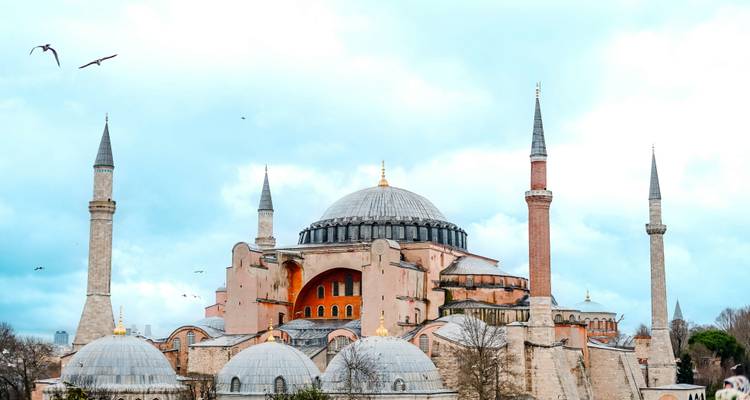 La Hagia Sophia con sus cuatro esbeltos minaretes se alza majestuosamente contra un cielo azul brillante con algunas aves que vuelan.