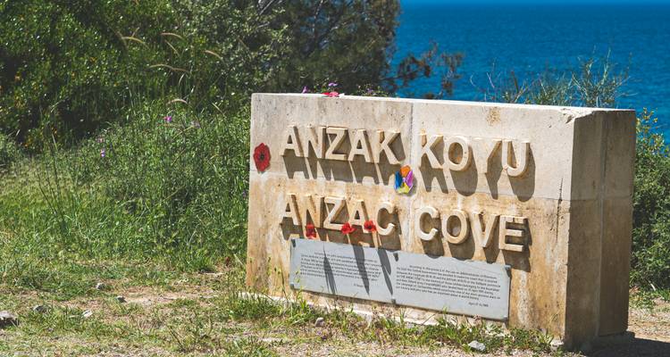 Memorial de piedra que dice "ANZAK KOYU / ANZAC COVE" adornado con amapolas, con vista a las aguas azul brillante del Egeo.