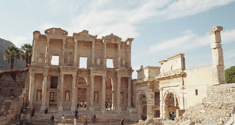 La gran fachada de la Biblioteca de Celso en Éfeso con visitantes explorando las antiguas ruinas de mármol bajo un cielo parcialmente nublado.