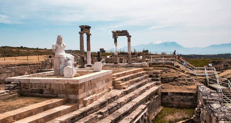 Ruinas grecorromanas dispersas con estatuas de mármol y columnas en una ladera; montañas cubiertas de nieve difusas en el fondo.