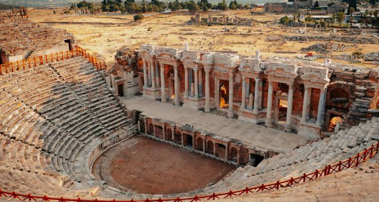 Vista panorámica de un anfiteatro romano antiguo con asientos de piedra escalonados y un escenario bien conservado en medio de un campo dorado.