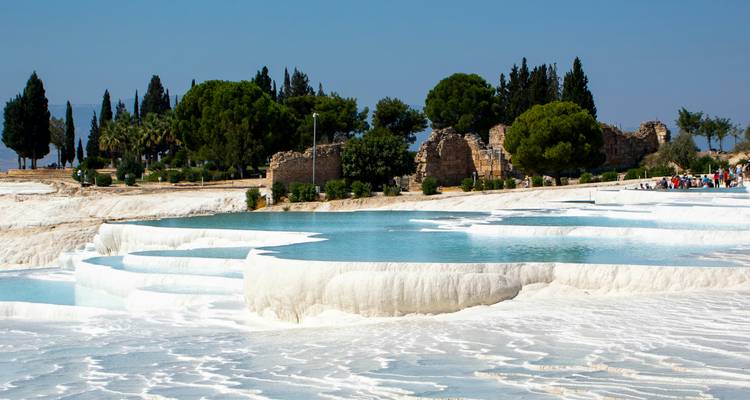 Terrazas de travertino blanco brillante llenas de agua turquesa en Pamukkale con árboles verdes y ruinas en la distancia.