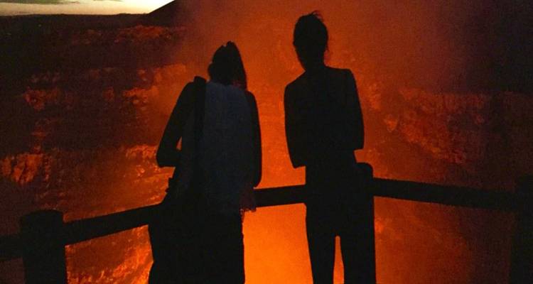Twee silhouetten leunen over een houten reling en kijken naar de vurige gloed van een actieve vulkaankrater in de nacht.