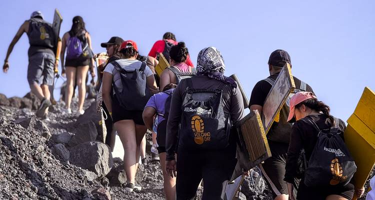 Een rij trekkers met planken beklimt een rotsachtige helling richting de vulkaantop op een zonnige dag.