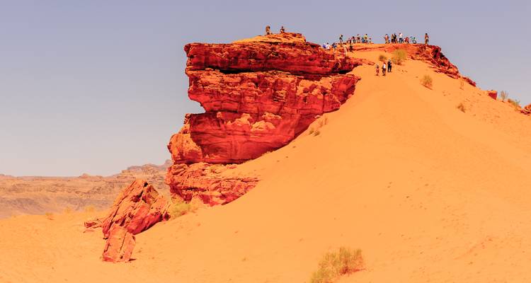 Besucher stehen auf einem roten Sandsteinaufschluss, der sich über die orangefarbenen Dünen des Wadi Rum erhebt.
