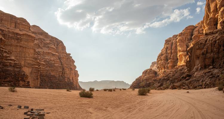 Breites Sandtal, flankiert von hoch aufragenden Sandsteinwänden unter einem teilweise bewölkten Himmel im Wadi Rum.
