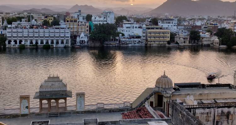 Ruhiger Sonnenuntergang über dem Pichola-See, der die weißen Fassaden der Udaipur-Gebäude mit den umgebenden Hügeln im Hintergrund widerspiegelt.