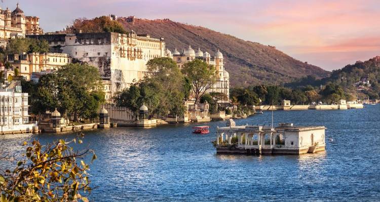 Schlossartiger Stadtpalast-Komplex, der sich zur goldenen Stunde über die blauen Wasser des Pichola-Sees erhebt, mit sanften Hügeln dahinter.