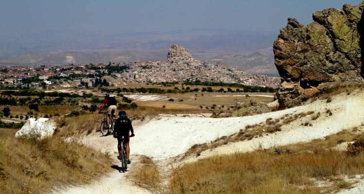 Cyclistes descendant un sentier de terre blanche vers une vue panoramique du village d'Uchisar et du paysage volcanique en Cappadoce.