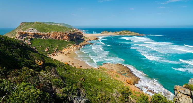 Dramatic viewpoint over turquoise bay, golden sand and rugged green peninsula with crashing waves.