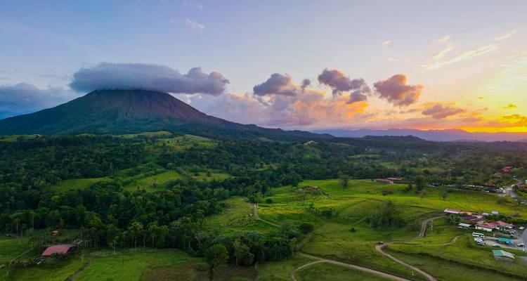 Vue de drone du volcan Arenal coiffé de nuages au-dessus de la campagne verdoyante vallonnée au coucher du soleil.
