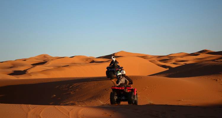 Two riders drive quad bikes up golden dunes under a clear desert sky in Merzouga.