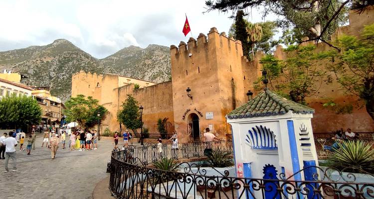 Place de la Kasbah de Chefchaouen avec des murs de forteresse ocre, un arrière-plan montagneux et des visiteurs.