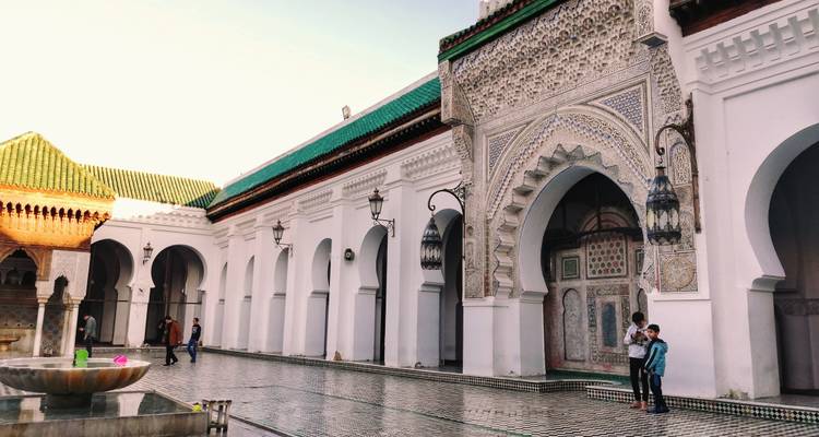 Des personnes se tiennent dans la cour carrelée ornementée d'une mosquée marocaine avec de grandes arches.