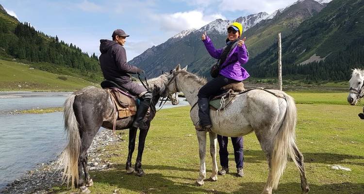 Two riders on horseback smile beside a mountain river with snowy peaks in the distance.