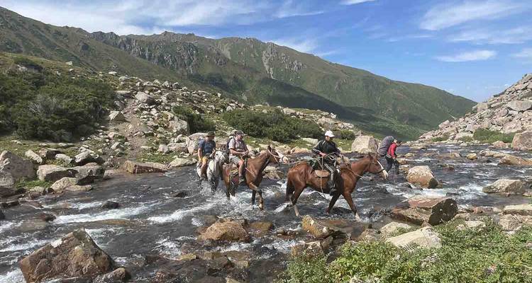 Group of riders guide their horses across a rocky mountain stream beneath a blue sky.