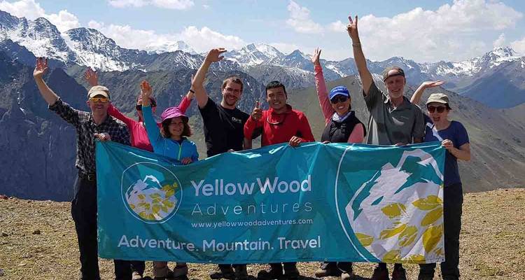 Happy trekking group holds a branded banner on a windy mountain ridge with snowy peaks behind.